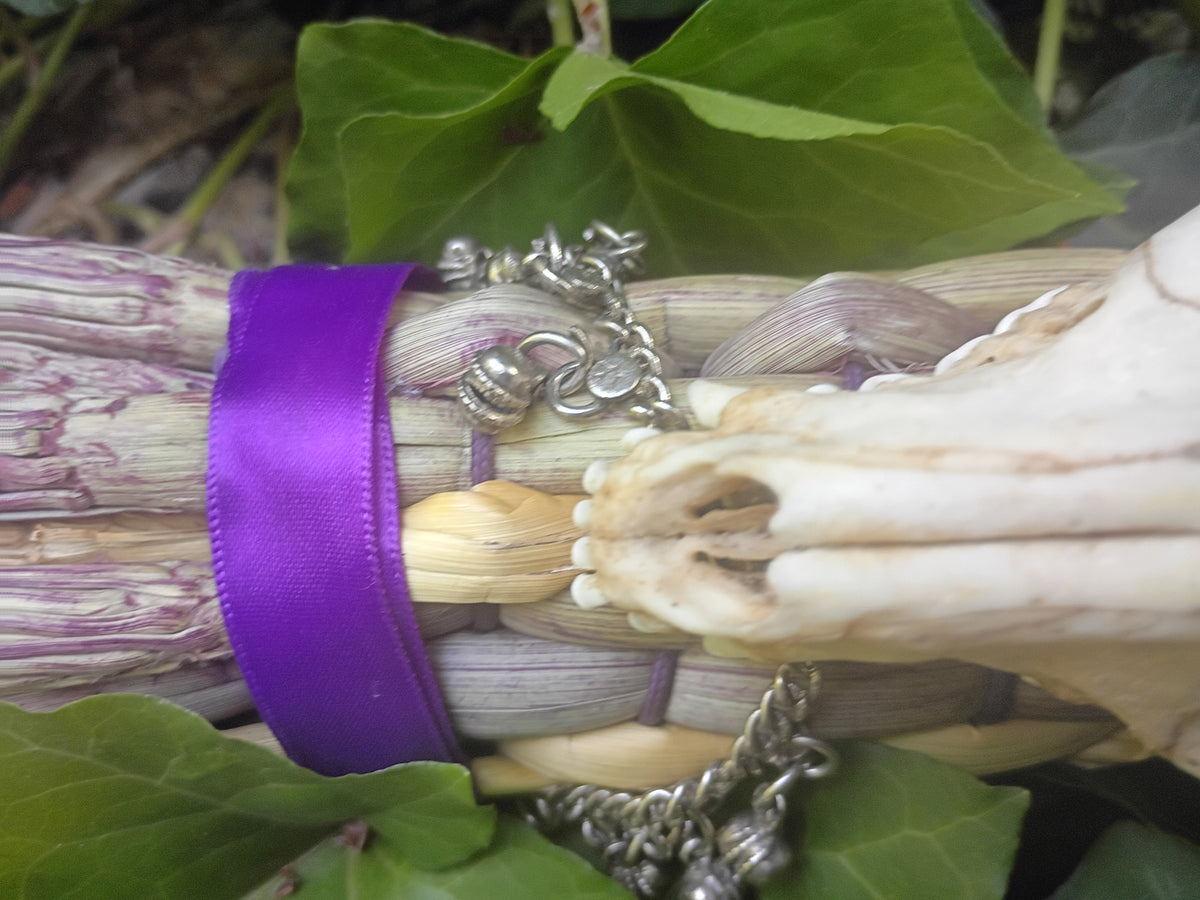A close-up of a silver charm bells wrapped around broom weave with a purple ribbon, set against green leaves.