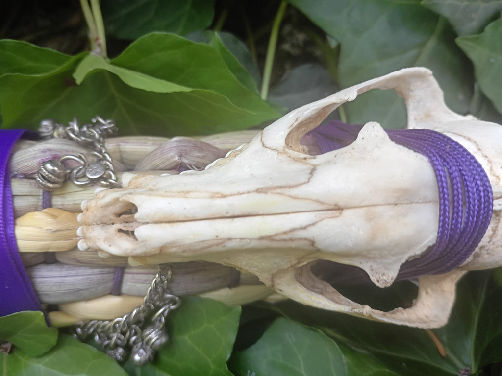 A close-up of a broom weave wrapped with purple cords, featuring a small animal skull placed on top, silver chain accents, and green leaves in the background.