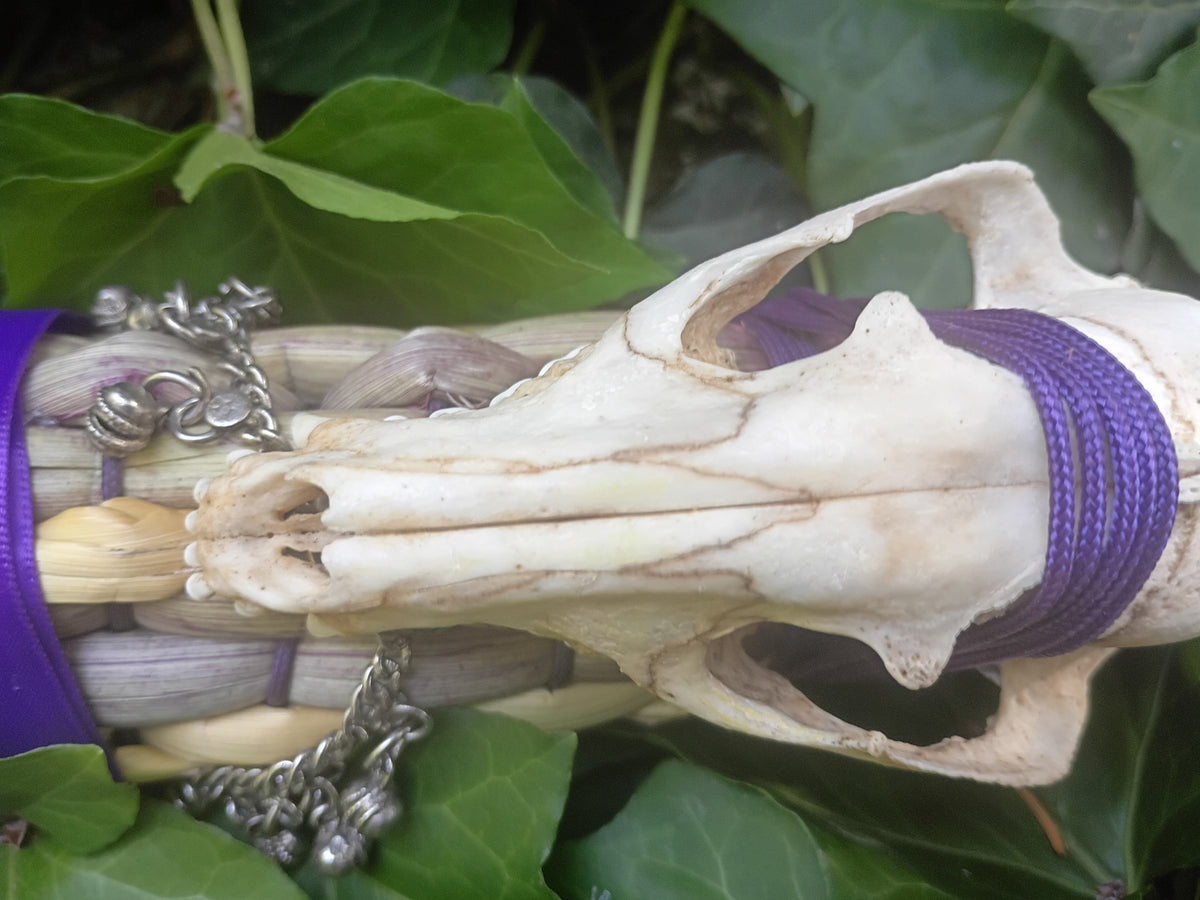 A close-up of a broom weave wrapped with purple cords, featuring a small animal skull placed on top, silver chain accents, and green leaves in the background.