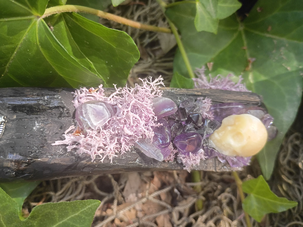 A close-up of the tip of the broom handle adorned with purple amethyst stones, light purple moss and a cream calcite stone skull set against a background of green ivy leaves.