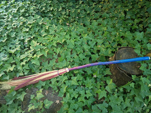 A long, driftwood broom handle with a blue and purple-to-red ombre pattern with natural and red hand dyed broomcorn bristles. Woven with Appalachian weave and purple cord. Tip of broom decorated with a cabochon style crystal. Broom is resting on a bed of ivy leaves.