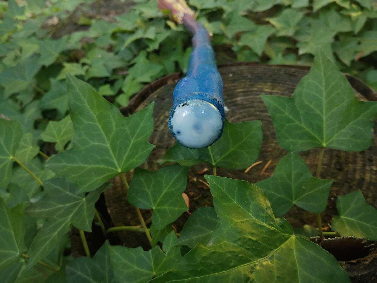 Tip of broom handle capped with a moss agate cabochon style stone, nestled among green ivy leaves outdoors.