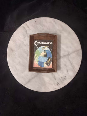 A rectangular dark stained wooden rolling tray with handles and laminated artwork sits on a round white marble tabletop. Illustration depicts a 1920s era flapper girl with short black hair, a teal cloche cap topped with a peacock feather, teal dress and ice blue fur shawl. The lady is smoking, has her head tilted up and blowing her smoke into the air against a black and pink background. In the bottom right corner is a picture of a pack of Conquistador rolling papers.