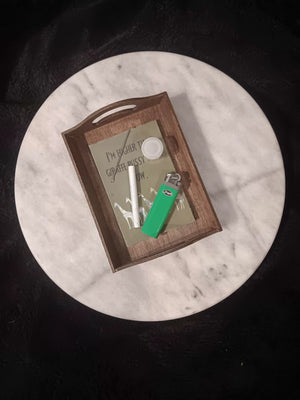 A rectangular dark stained wooden rolling tray with handles and laminated artwork sits on a round white marble tabletop. The  illustration shows a humorous message reading 'I'M HIGHER THAN GIRAFFE PUSSY RIGHT NOW.' above five colorful patterned giraffe illustrations on an olive background. Tray contains a doobie, a small container of wax, a dabber tool, and a bright green lighter.