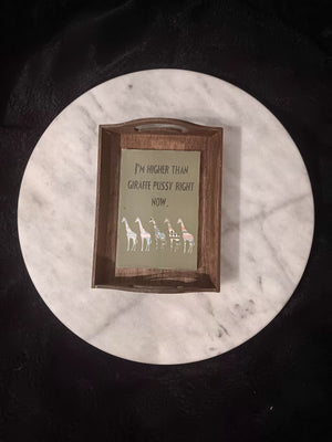 A rectangular dark stained wooden rolling tray with handles and laminated artwork sits on a round white marble tabletop. The  illustration shows a humorous message reading 'I'M HIGHER THAN GIRAFFE PUSSY RIGHT NOW.' above five colorful patterned giraffe illustrations on an olive background. 