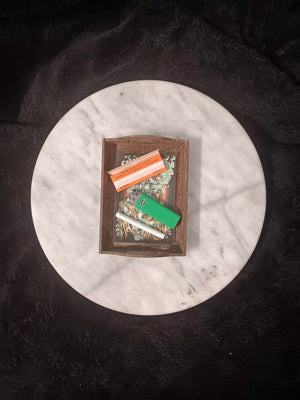 A rectangular dark stained wooden rolling tray with handles and laminated artwork sits on a round white marble tabletop. The  illustration shows a gray and silver shaded Shaman wearing a ram's head, hitting a skull shaped bong with smoke surrounding him on a red and black background. Tray contains a doobie,  a pack of rolling papers and a bright green lighter.